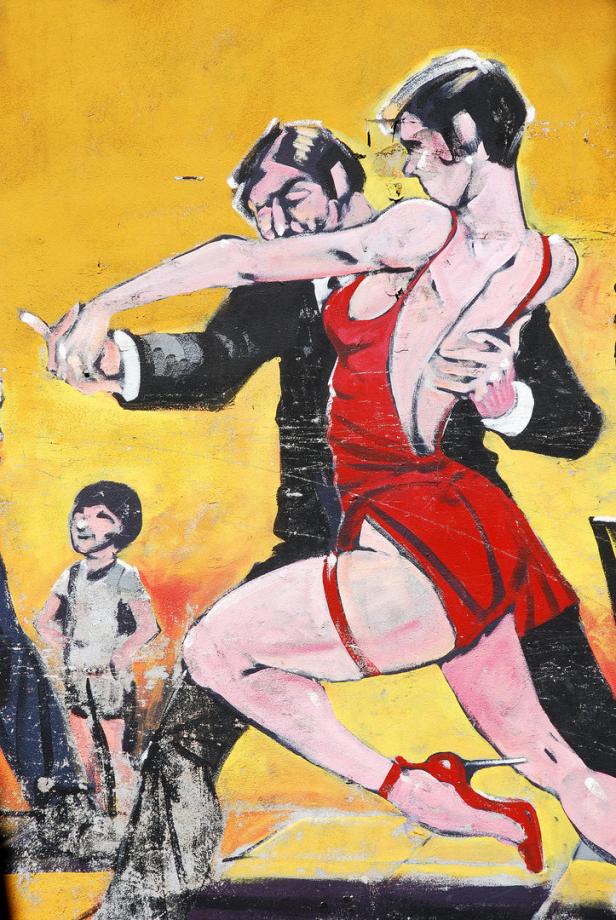 Ein Paar tanzt Tango vor einer gelben Wand mit einem Kind im Hintergrund.