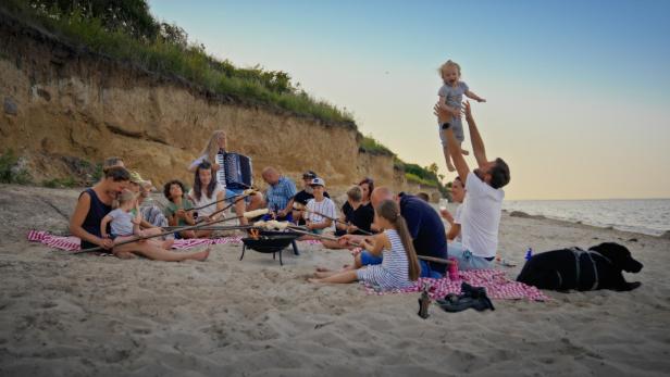 Eine Gruppe von Menschen sitzt am Strand und grillt Stockbrot, während ein Mann ein Kind in die Luft hebt.