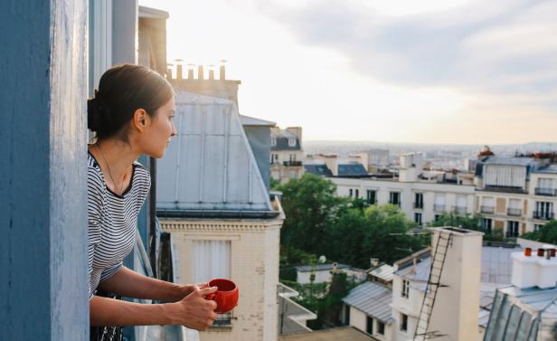 Eine Frau blickt mit einer Tasse in der Hand von einem Pariser Balkon auf die Stadt.