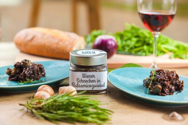 Ein Glas „Wiener Schnecken in Balsamicozwiebel“ steht neben zwei Tellern mit Schnecken und Zwiebeln.