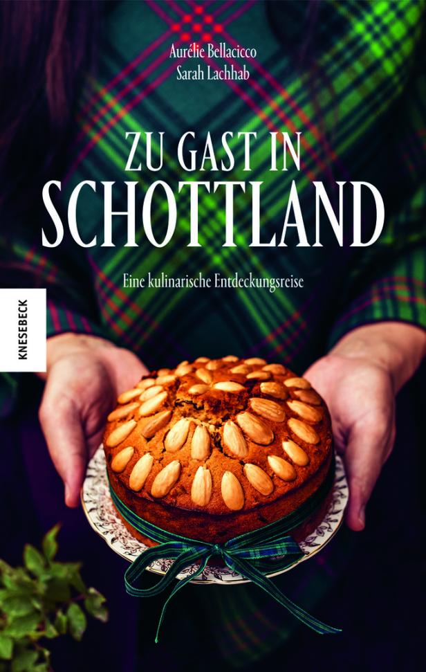 Ein Buchcover von „Zu Gast in Schottland“ mit einem Mandelkuchen, der von Händen gehalten wird.