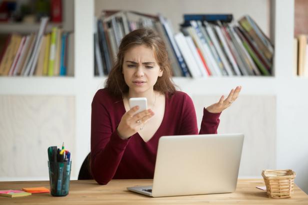 Eine Frau sitzt vor einem Laptop und blickt verwirrt auf ihr Smartphone.