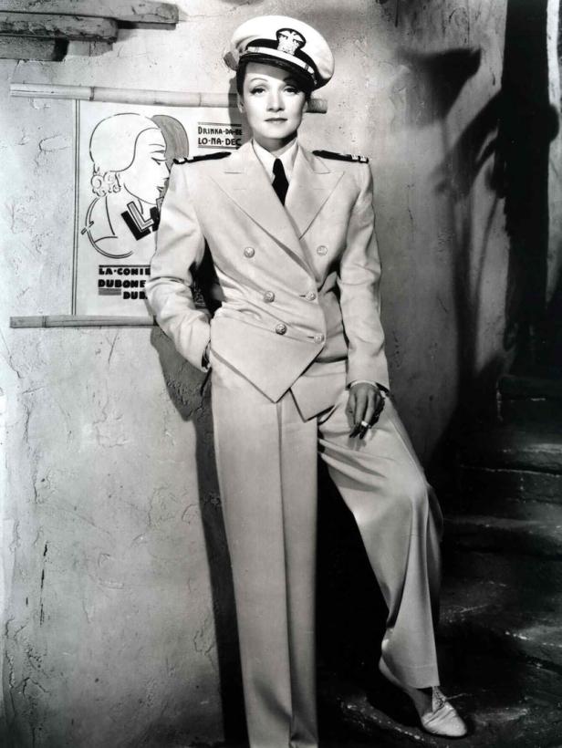 Marlene Dietrich in Uniform mit Zigarette, posierend vor einem Werbeplakat.