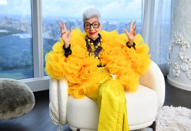 Iris Apfel posiert in einem gelben Outfit in einem Sessel vor einem Fenster.