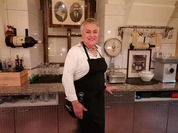 Eine Frau mit Schürze steht in einer Küche mit alten Fotos und Küchengeräten.