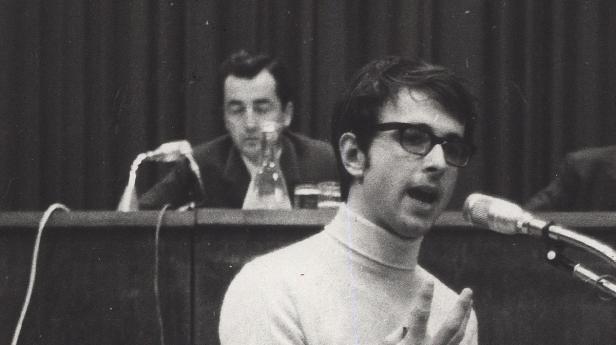 Ein junger Mann mit Brille spricht an einem Mikrofon, eine weitere Person im Hintergrund.