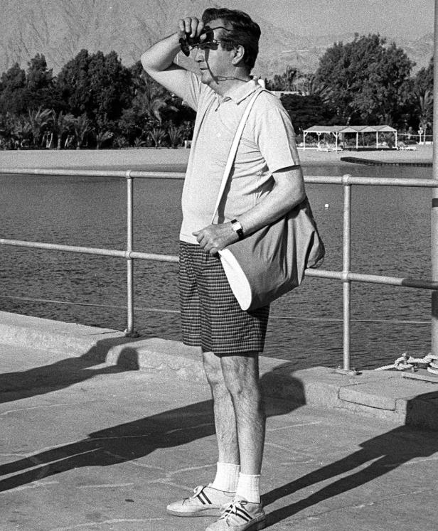 Ein Mann mit Shorts, Poloshirt und Tasche steht am Wasser und hält eine Sonnenbrille hoch.