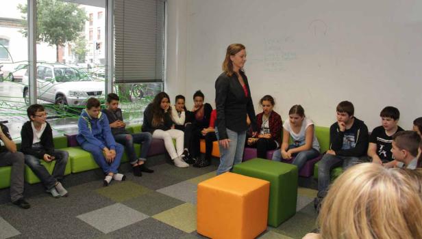 Eine Lehrerin steht vor einer Gruppe von Schülern, die auf bunten Sitzgelegenheiten sitzen.