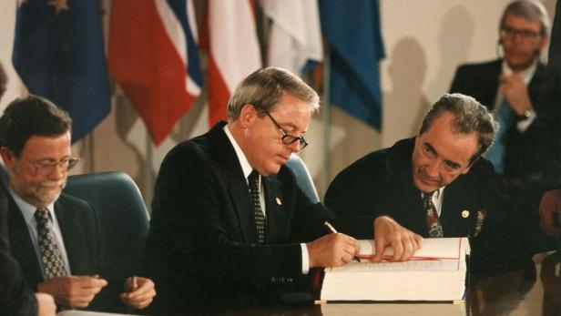 Drei Männer in Anzügen unterzeichnen ein Dokument vor Flaggen.