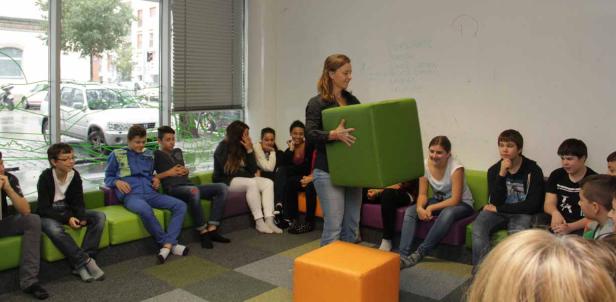 Eine Frau hält einen grünen Würfel vor einer Gruppe von Jugendlichen in einem Klassenzimmer.