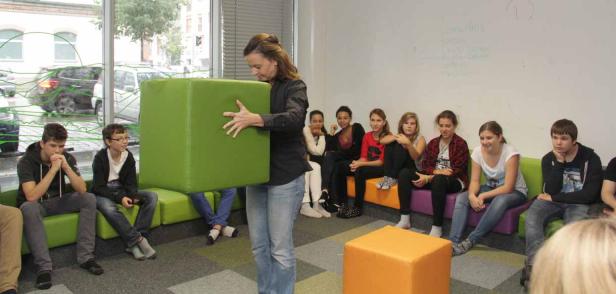 Eine Frau trägt einen großen grünen Würfel vor einer Gruppe von Jugendlichen.