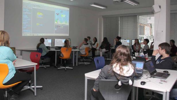 Schüler arbeiten in einem Klassenzimmer an Computern mit einer Projektion von Scratch-Code an der Wand.