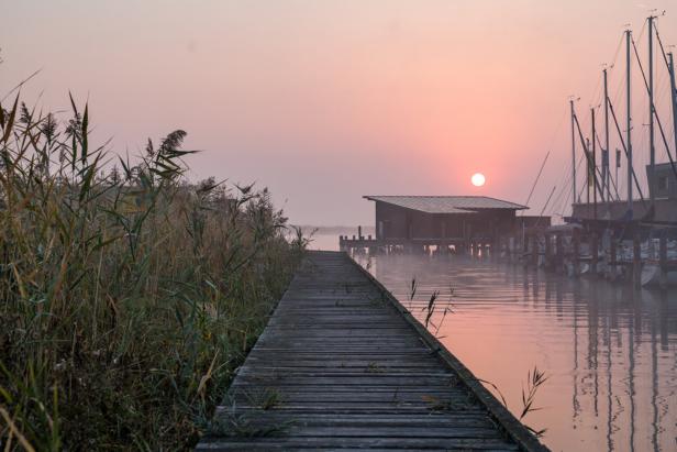 Ein Holzsteg führt zu einem kleinen Haus am See im Morgennebel.