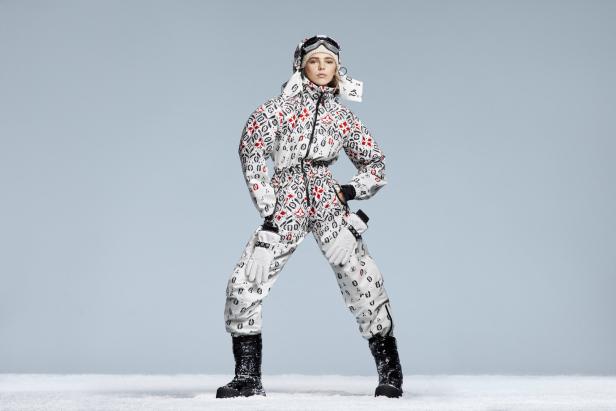 Eine Frau in einem gemusterten Skianzug, Handschuhen und Stiefeln steht im Schnee.