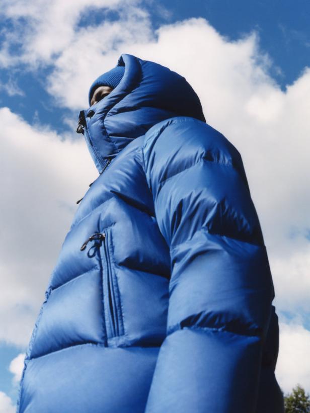 Eine Person mit blauer Mütze und Daunenjacke steht vor einem Himmel mit Wolken.