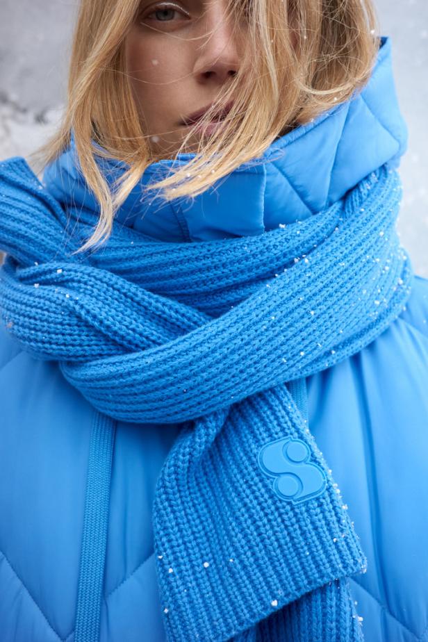 Eine Frau mit blonden Haaren trägt einen blauen Schal und eine passende Jacke im Schnee.