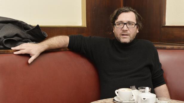 Ein Mann mit Brille sitzt in einem Café auf einer roten Bank und trinkt Kaffee.