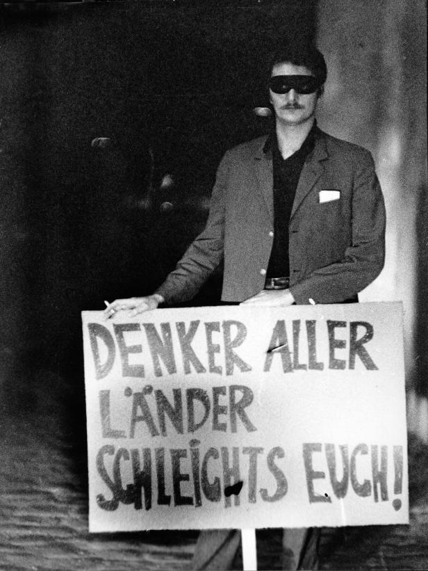 Ein Mann mit Sonnenbrille hält ein Schild mit der Aufschrift „Denker aller Länder, schleichts euch!“.