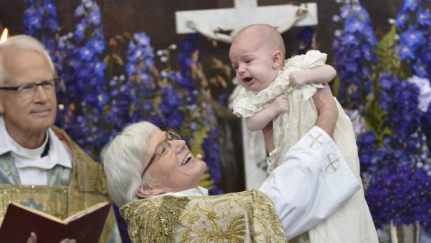 Ein weinendes Baby wird während einer Taufe von einer Pfarrerin hochgehalten.