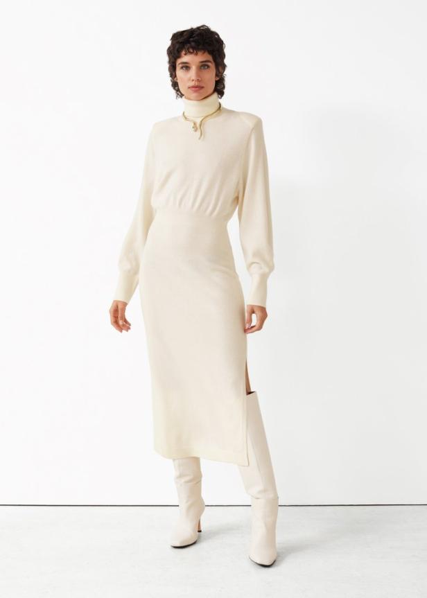 Eine Frau in einem cremefarbenen Rollkragenkleid und passenden Stiefeln posiert vor einem weißen Hintergrund.