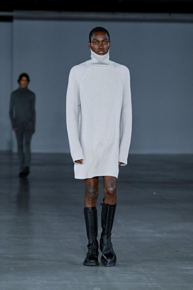 Ein Model präsentiert ein helles Strickkleid mit Rollkragen und hohe Schnürstiefel auf dem Laufsteg.