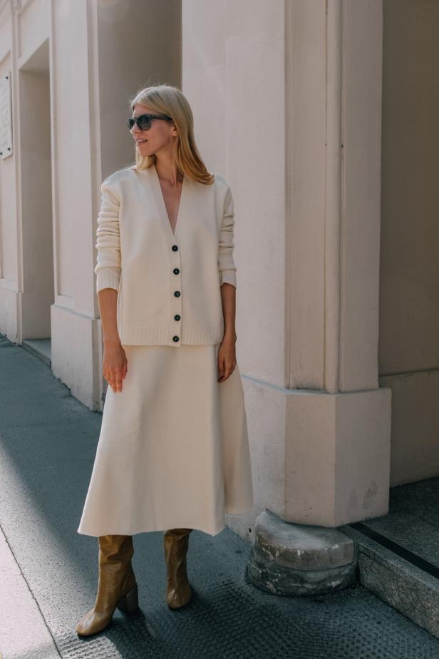 Eine blonde Frau trägt einen cremefarbenen Cardigan, einen Rock und braune Stiefel.