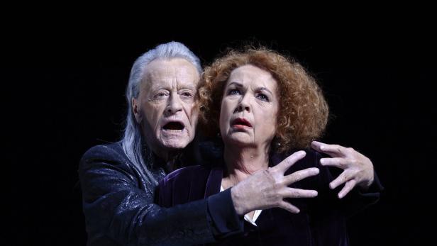 Ein älterer Mann mit langen, grauen Haaren umarmt eine Frau mit roten Locken, beide mit besorgten Gesichtsausdrücken.