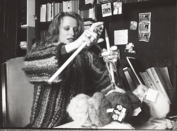 Eine Frau strickt mit dicken Stricknadeln vor einem Bücherregal.