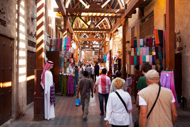 Menschen flanieren durch einen belebten Souk mit Textilien und traditioneller Architektur.