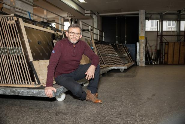 Ein Mann mit Brille sitzt auf einem Rollwagen in einer Lagerhalle.