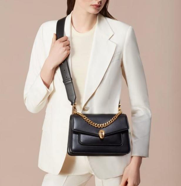 Eine Frau trägt eine schwarze Handtasche mit goldener Kette und einem weißen Hosenanzug.