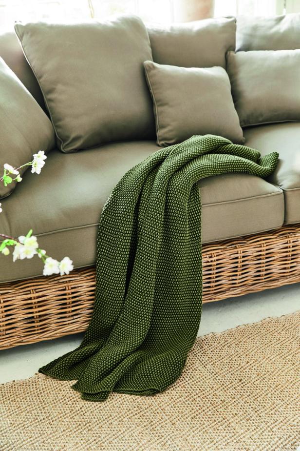 Eine grüne Strickdecke liegt auf einem beigen Sofa mit Rattankorb-Basis.