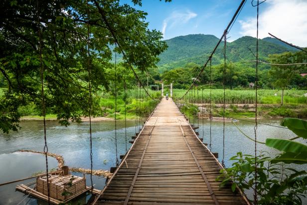 Eine Hängebrücke aus Bambus führt über einen Fluss in einer grünen Landschaft.
