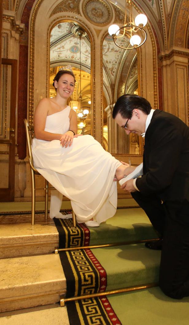Eine Frau im weißen Kleid und mit Tiara sitzt, während ein Mann ihr den Schuh anpasst.