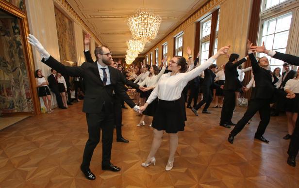 Eine Gruppe junger Leute tanzt in einem festlichen Saal mit Parkettboden und Kristalllüstern.