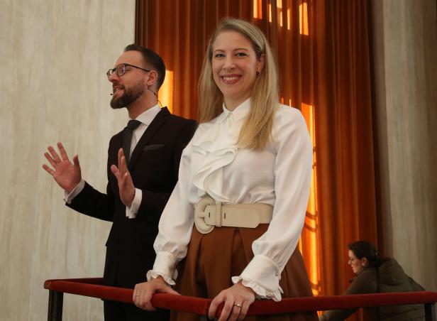 Eine Frau lächelt neben einem Mann mit Brille und Anzug auf einer Bühne.
