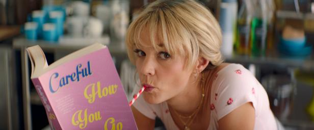 Eine blonde Frau trinkt mit einem Strohhalm und liest das Buch „Careful How You Go“.