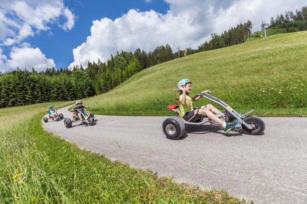 Drei Kinder fahren mit Bergrollern eine asphaltierte Straße hinunter.