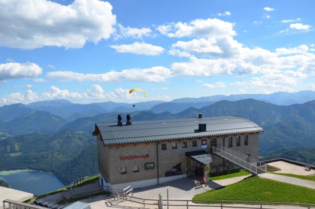 Das Terzerhaus thront vor einer Bergkulisse unter blauem Himmel, ein Gleitschirmflieger ist zu sehen.