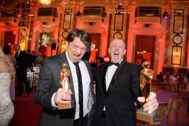 Zwei Männer im Smoking jubeln mit ihren Romy-Auszeichnungen in einem festlichen Saal.