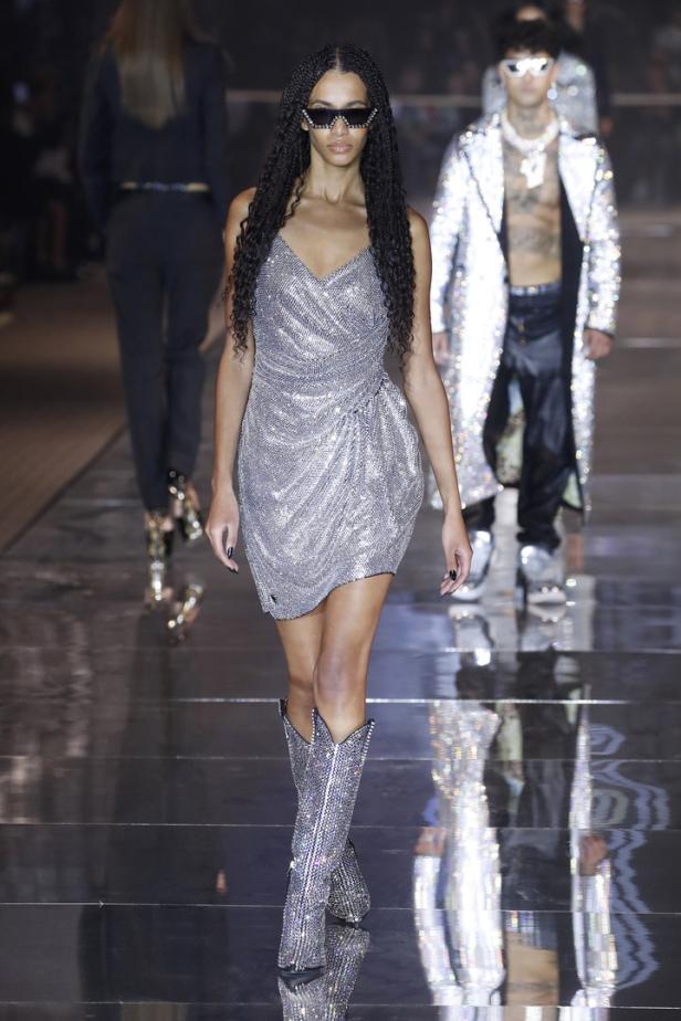 Eine Frau mit Sonnenbrille und glitzerndem Kleid und Stiefeln läuft über einen Laufsteg.
