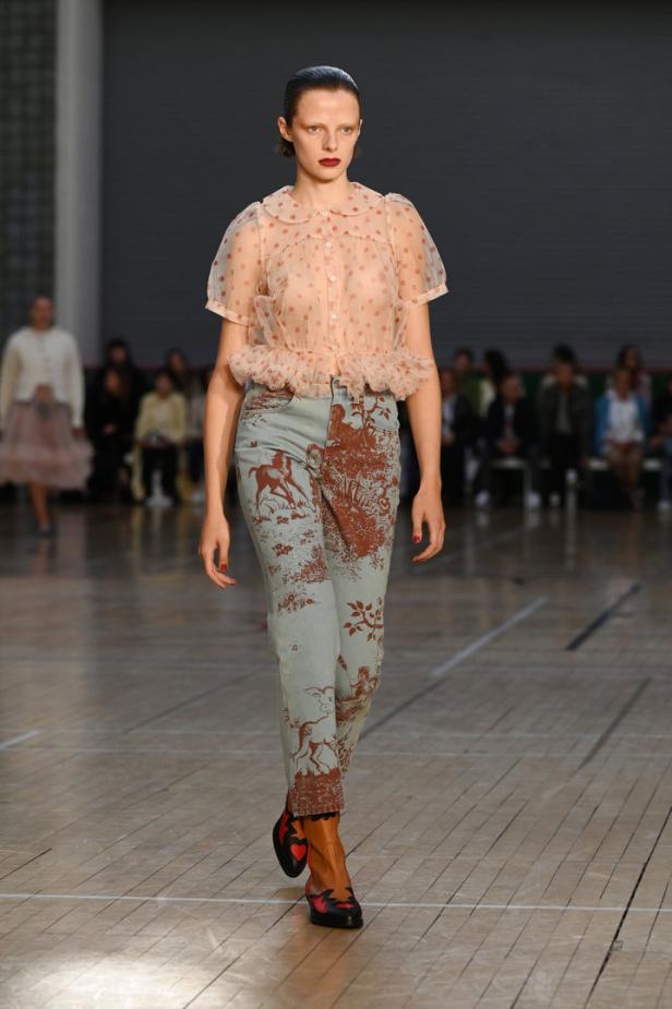 Eine Frau auf dem Laufsteg trägt eine transparente Bluse und bedruckte Jeans.