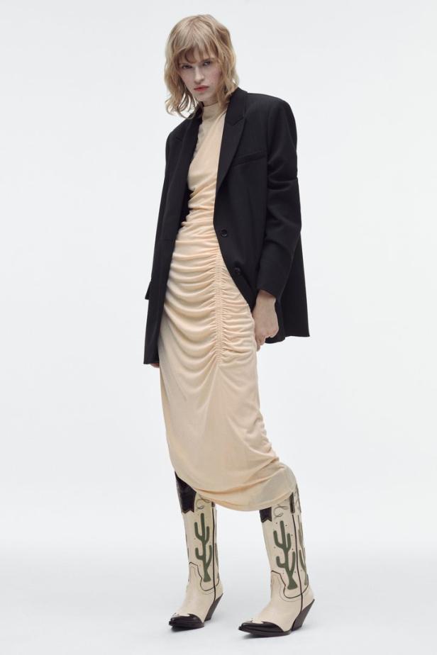 Eine Frau mit blonden Haaren trägt ein Kleid, einen Blazer und Cowboystiefel mit Kaktusmotiven.