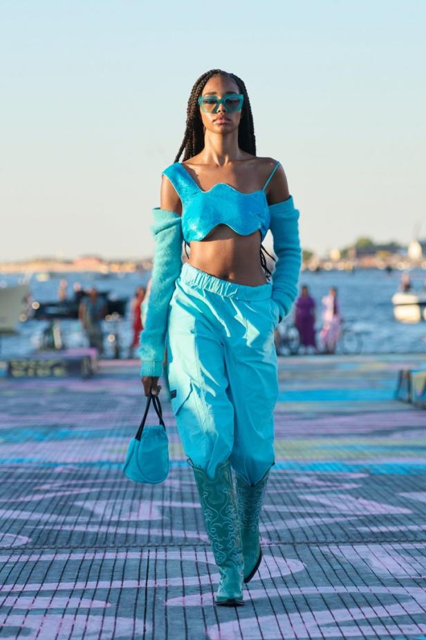 Eine Frau in einem türkisfarbenen Outfit läuft auf einem Laufsteg am Wasser.