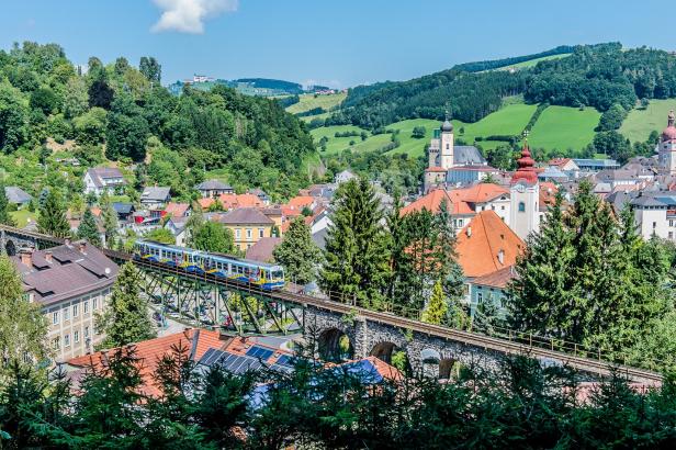 Ein Zug fährt über eine Brücke in einer Stadt, umgeben von Gebäuden und urbaner Landschaft.