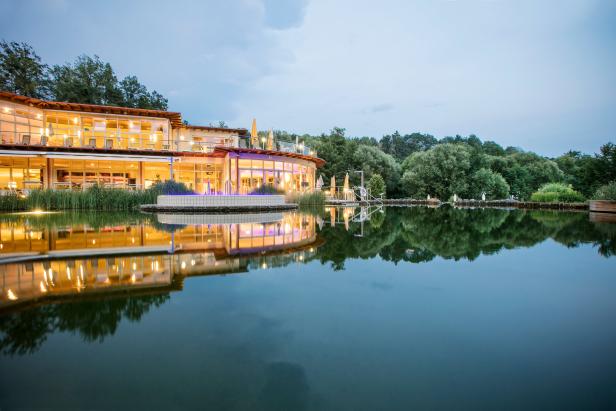 Ein modernes Gebäude mit vielen Fenstern spiegelt sich in einem ruhigen See.