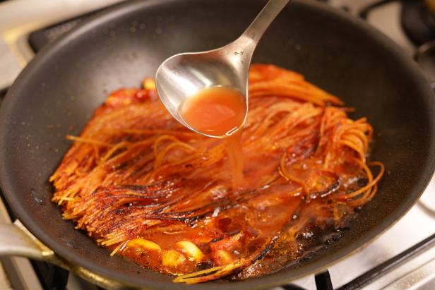 Mit einer Schöpfkelle wird Soße über gebratene Spaghetti in einer Pfanne gegossen.