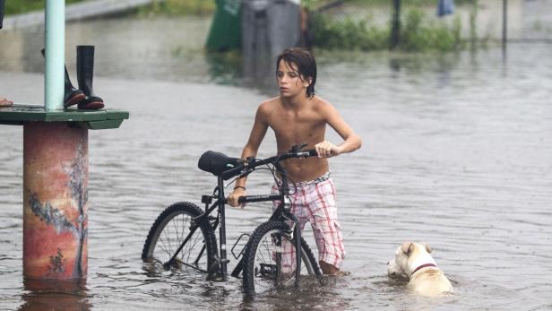 Ein Junge schiebt sein Fahrrad durch das knietiefe Hochwasser, begleitet von einem Hund.