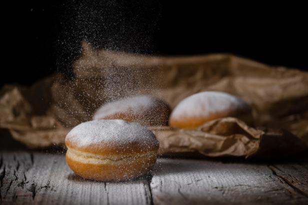 Drei Krapfen liegen auf einem Holztisch, während Puderzucker darüber gestreut wird.