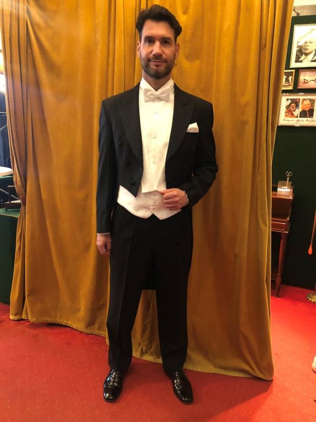 Ein Mann in formeller Abendgarderobe mit Fliege und Kummerbund steht vor einem goldenen Vorhang.